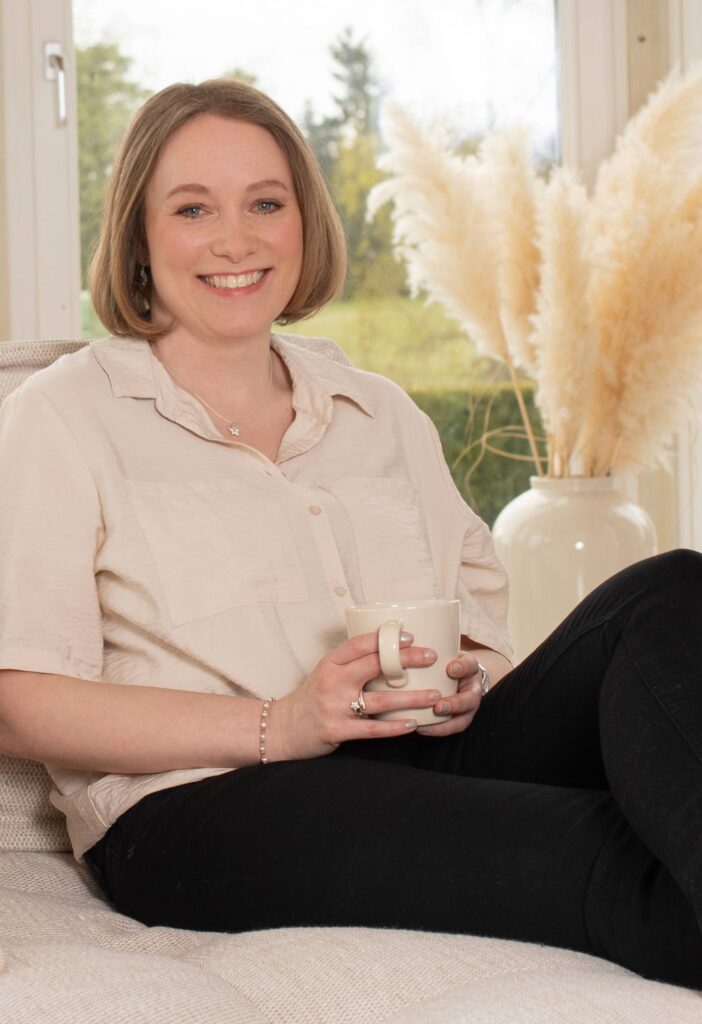 Carina Göckeler sitzt mit einer Tasse auf einem Sofa und lächelt in die Kamera.