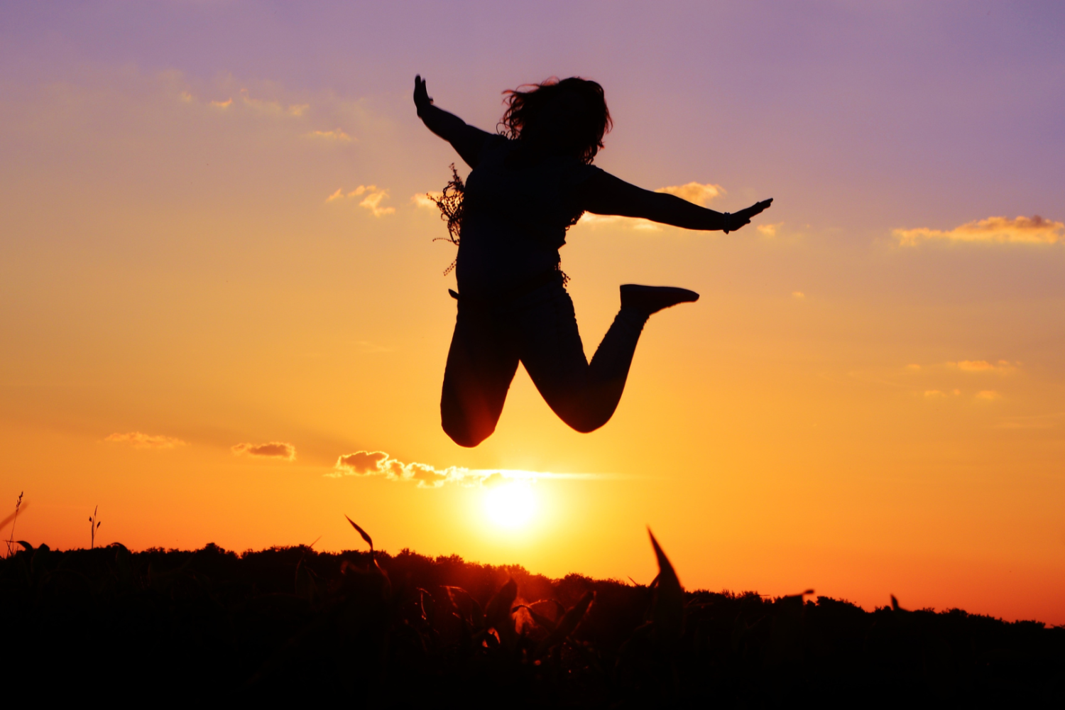 Silhouette einer Frau, die mit ausgebreiteten Armen vor einem Sonnenuntergang in die Luft springt
