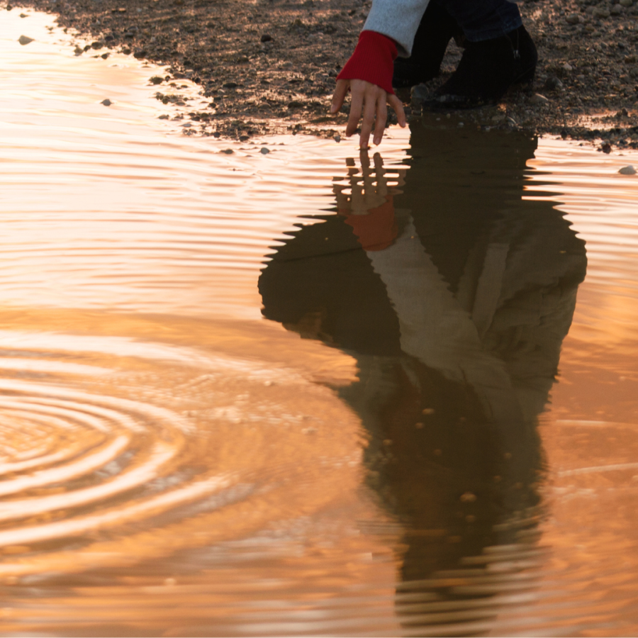 Eine Hand berührt eine Wasseroberfläche im goldenen Licht, darunter spiegelt sich die Person, die sie berührt