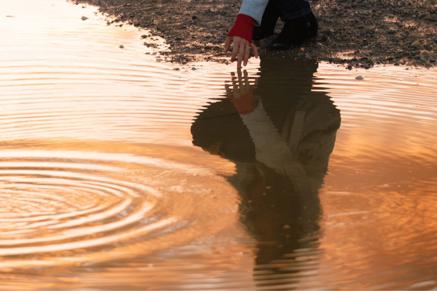 Eine Hand berührt eine Wasseroberfläche im goldenen Licht, darunter spiegelt sich die Person, die sie berührt
