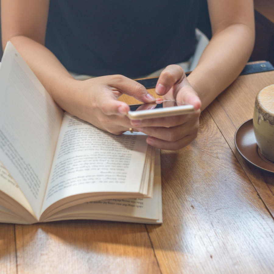 Eine Person sitzt im Café an einem Holztisch mit Cappuccino und hält abgelenkt ein Smartphone über einem aufgeschlagenen Buch, passend zum Thema schnelles Konsumieren.