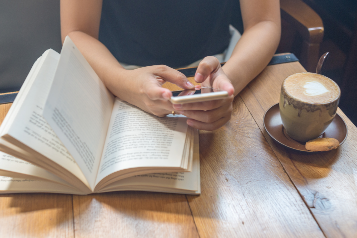 Eine Person sitzt im Café an einem Holztisch mit Cappuccino und hält abgelenkt ein Smartphone über einem aufgeschlagenen Buch, passend zum Thema schnelles Konsumieren.