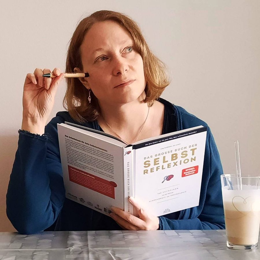 Carina sitzt am Tisch, hält das Buch ‚Das große Buch der Selbstreflexion‘ in der Hand, den Stift am Kopf, und schaut nachdenklich zur Seite. Neben ihr steht ein Latte macchiato.