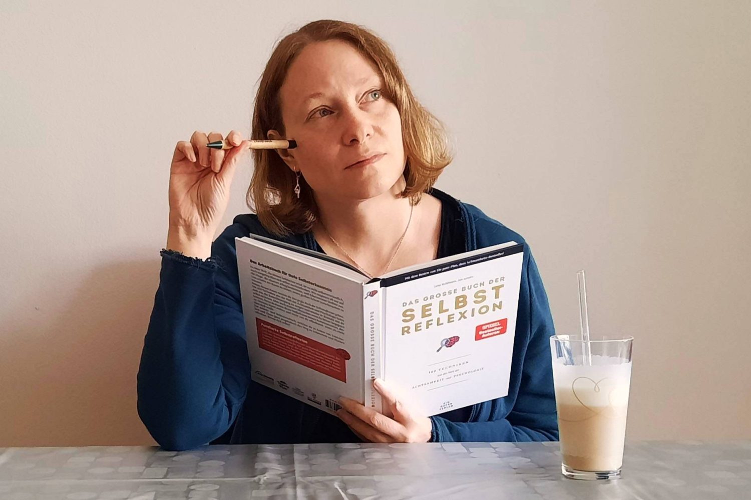Carina sitzt am Tisch, hält das Buch ‚Das große Buch der Selbstreflexion‘ in der Hand, den Stift am Kopf, und schaut nachdenklich zur Seite. Neben ihr steht ein Latte macchiato.